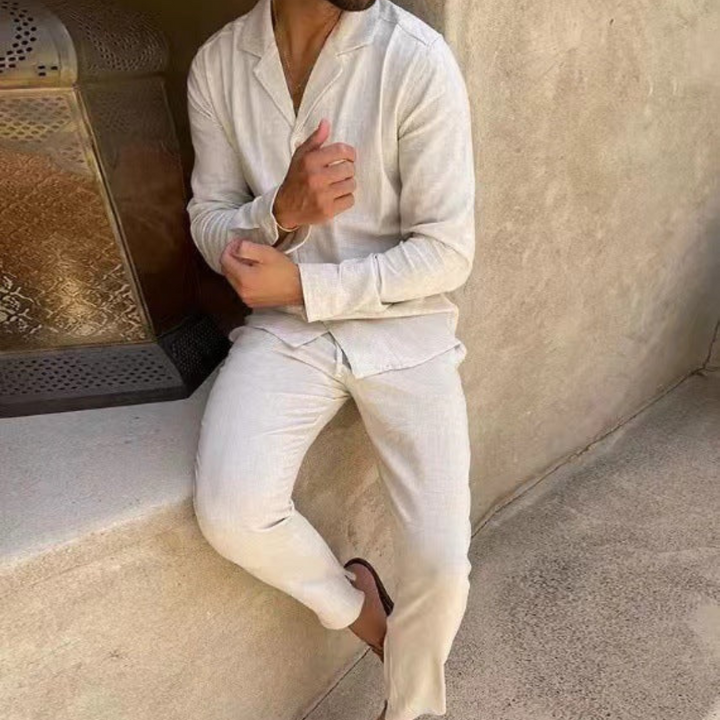 Mann in beiger Leinenkleidung lehnt an einer Wand, trägt Sandalen. Sommermode, lässiger Stil, entspannte Atmosphäre.