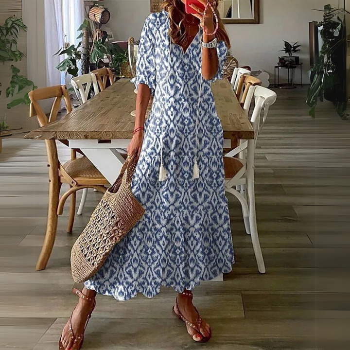 Frau in blau-weißem Boho-Kleid mit Ikat-Muster, hält eine Strohtasche, steht in modernem Esszimmer mit Holztisch und Pflanzen.