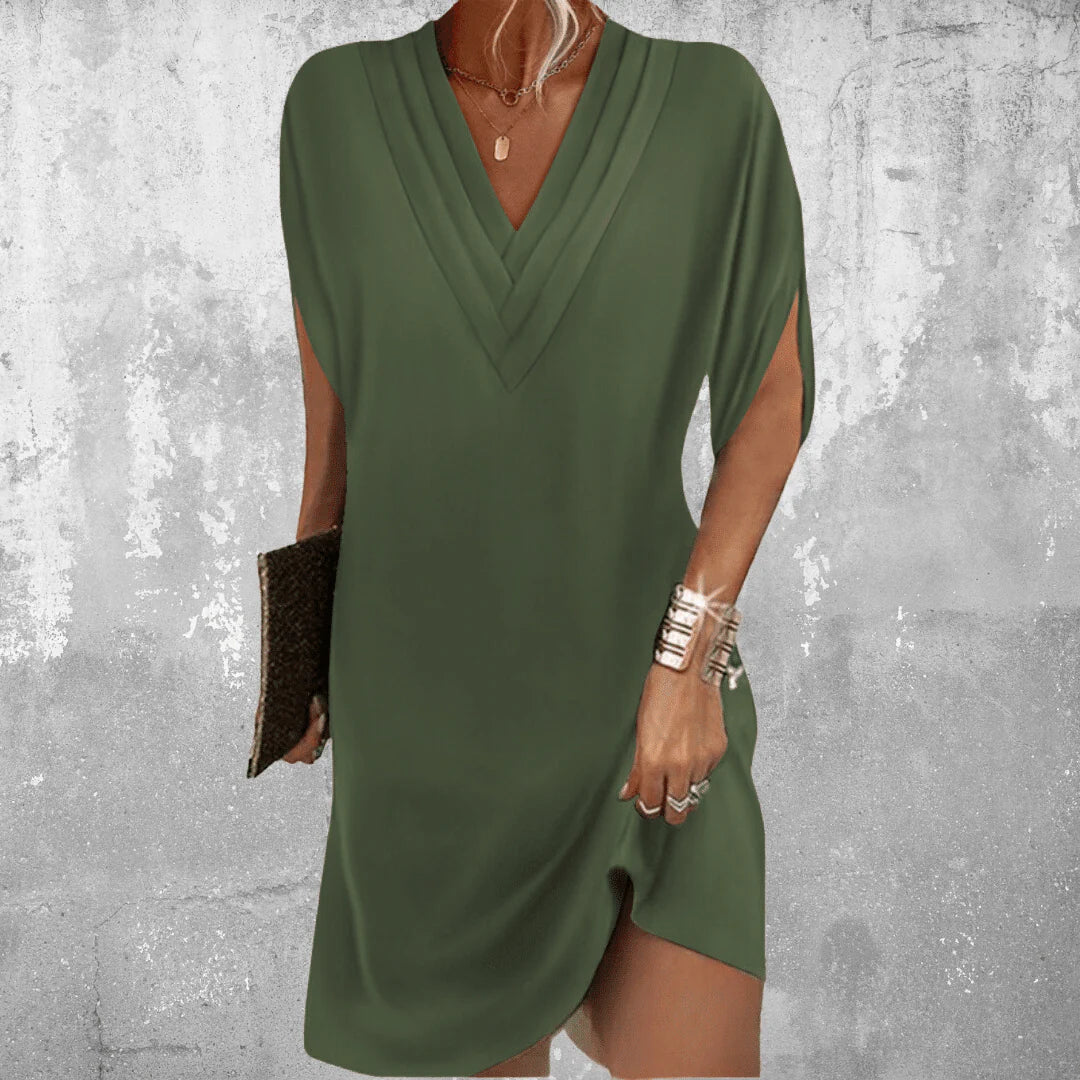 Frau in olivgrünem V-Ausschnitt-Kleid, hält schwarze Clutch, trägt silberne Armreifen, vor grauer Wand. Elegantes Sommeroutfit, modisch, stilvoll.