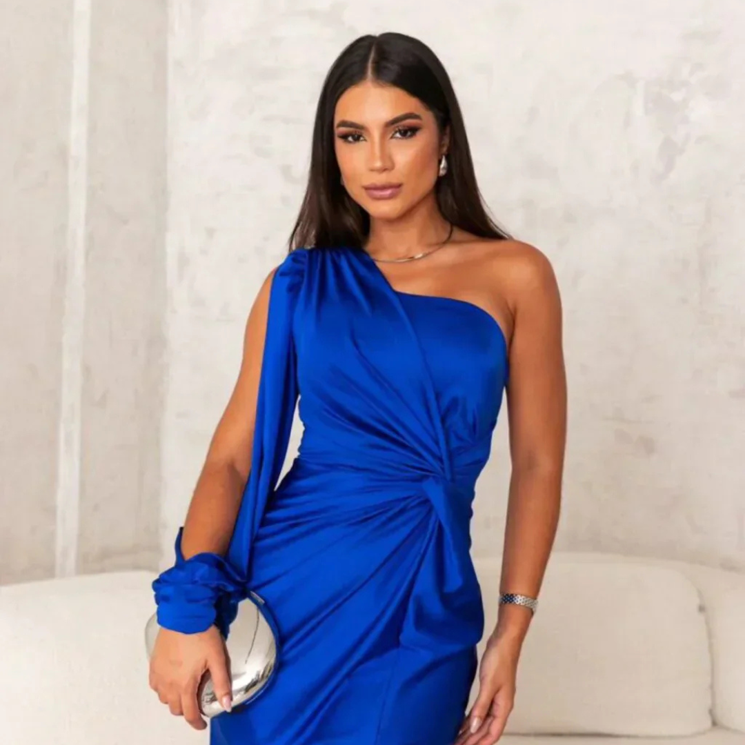 Eine Frau in einem eleganten, blauen, schulterfreien Kleid posiert vor einer neutralen Wand. Sie hält eine silberne Clutch und trägt dezente Ohrringe.