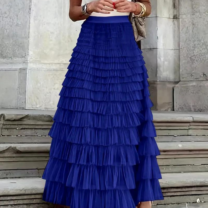Frau in blauem, mehrlagigem Rüschenrock vor steinernem Hintergrund. Eleganter, modischer Look, ideal für Sommermode und festliche Anlässe.
