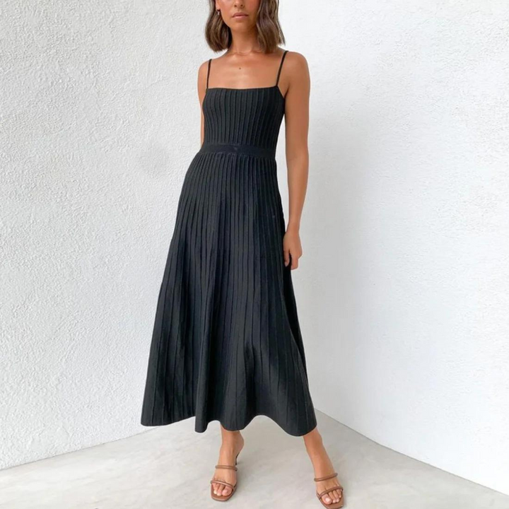 Frau in schwarzem, ärmellosem, plissiertem Maxikleid vor weißer Wand, trägt Sandalen. Elegantes Sommeroutfit, Mode, Damenbekleidung.