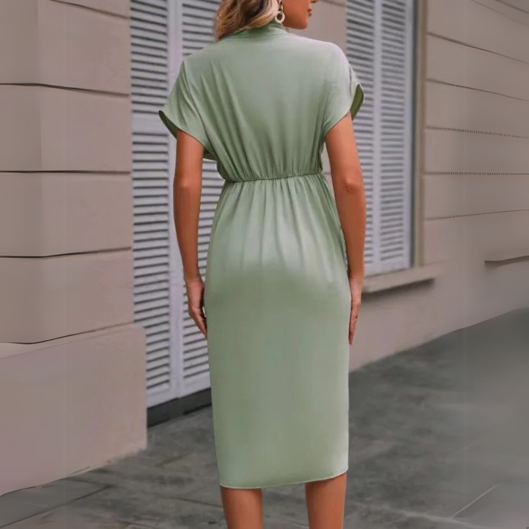 Frau in grünem, knielangem Kleid mit kurzen Ärmeln, steht vor einem Gebäude mit Fensterläden. Elegantes Sommeroutfit, Mode, Damenbekleidung.
