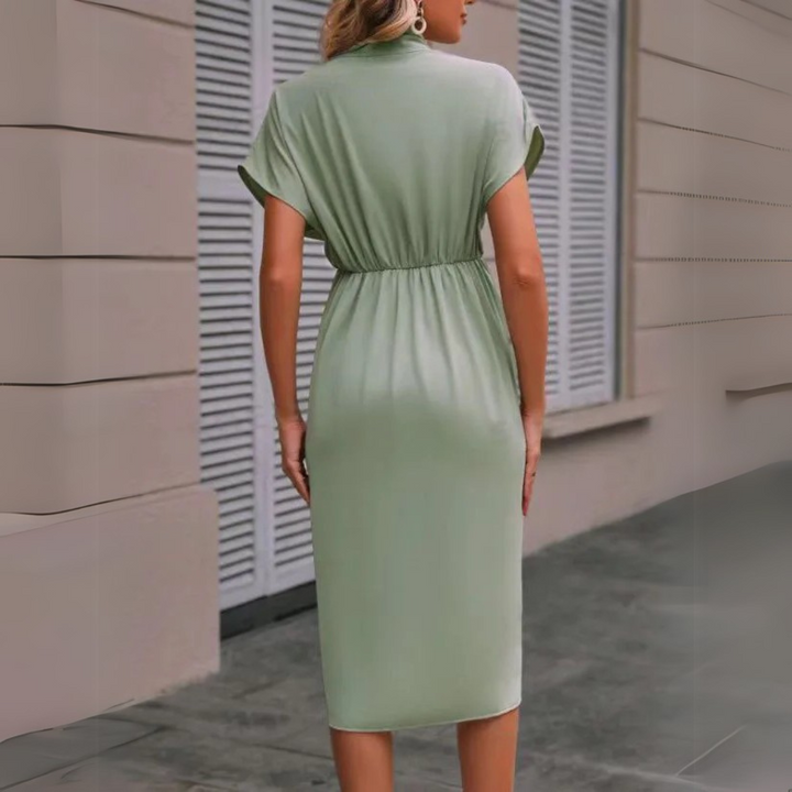 Frau in grünem, knielangem Kleid mit kurzen Ärmeln, steht vor einem Gebäude mit Fensterläden. Elegantes Sommeroutfit, Mode, Damenbekleidung.