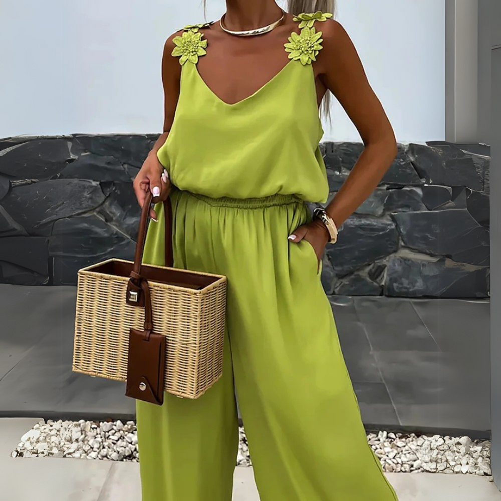 Frau in grünem Jumpsuit mit Blumenapplikationen, hält eine Korbtasche. Sommermode, elegantes Outfit, stilvolle Accessoires.