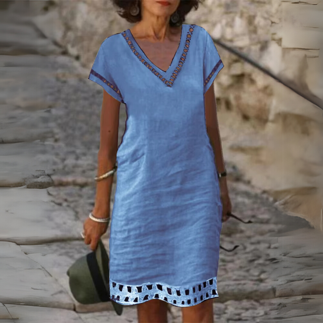 Frau in blauem Sommerkleid mit V-Ausschnitt und Stickereien, hält Hut in der Hand, steht auf gepflastertem Weg. Modetrend, Sommermode, Damenkleidung.