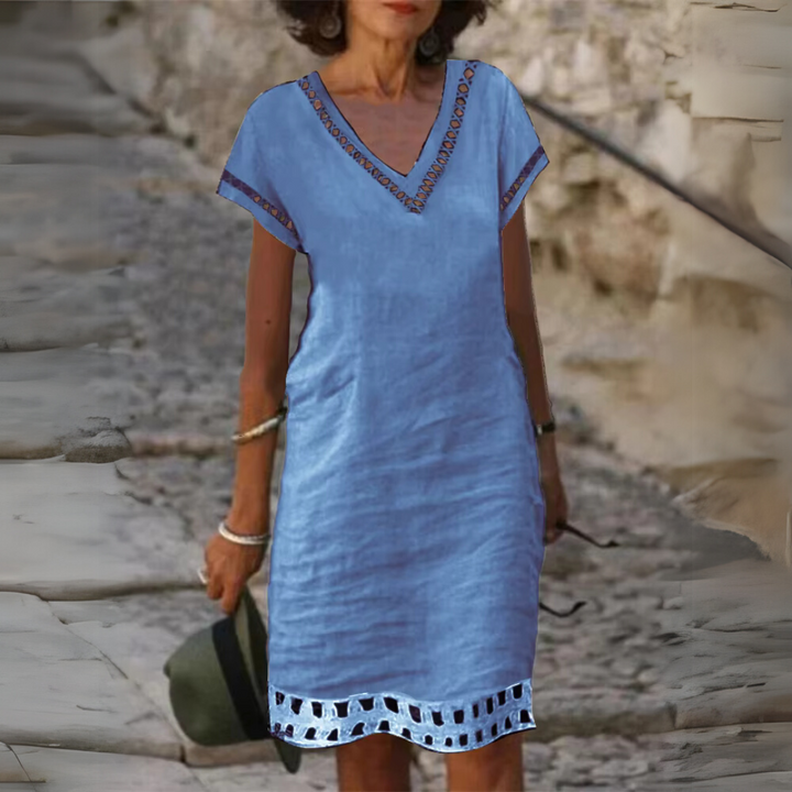 Frau in blauem Sommerkleid mit V-Ausschnitt und Stickereien, hält Hut in der Hand, steht auf gepflastertem Weg. Modetrend, Sommermode, Damenkleidung.