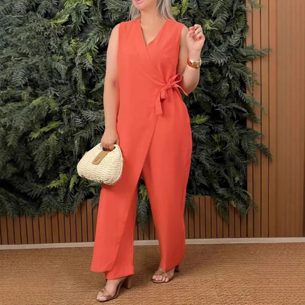 Frau in rotem Jumpsuit vor grüner Pflanzenwand, hält weiße Handtasche, trägt Sandalen. Mode, Sommeroutfit, Damenbekleidung, stilvoll.