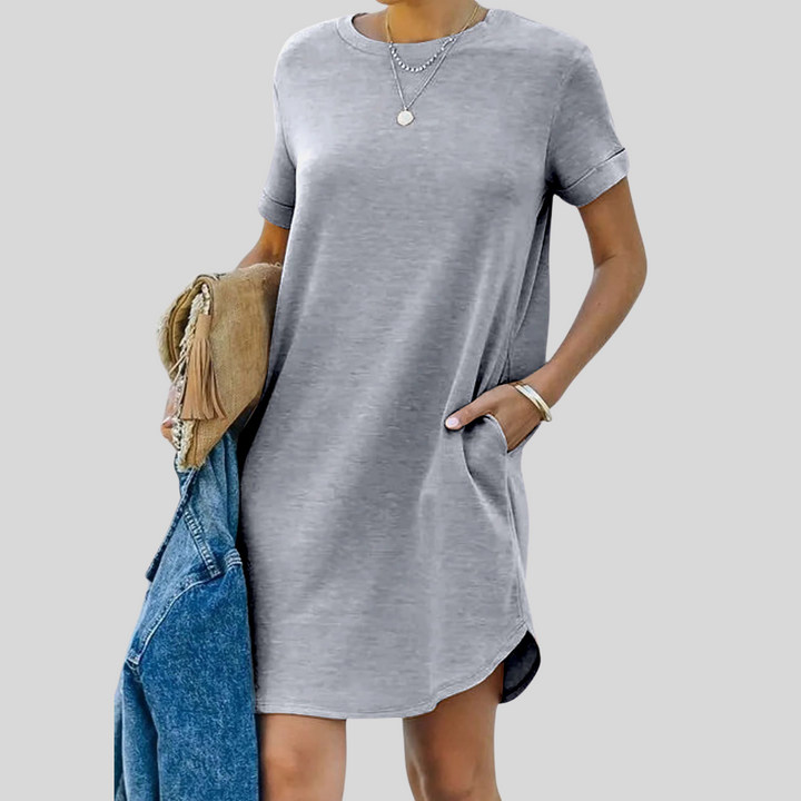 Frau in grauem T-Shirt-Kleid mit Taschen, hält blaue Jeansjacke und braune Tasche. Modetrend, lässiger Stil, Sommeroutfit.