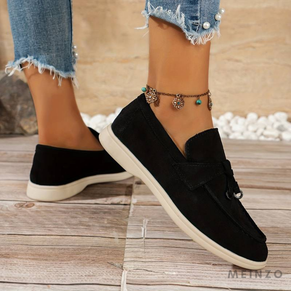 Damenfüße in schwarzen Mokassins auf Holzboden, Jeans mit Perlenbesatz, Fußkettchen mit Anhängern. Modische Schuhe, Freizeitlook, Sommerstil.