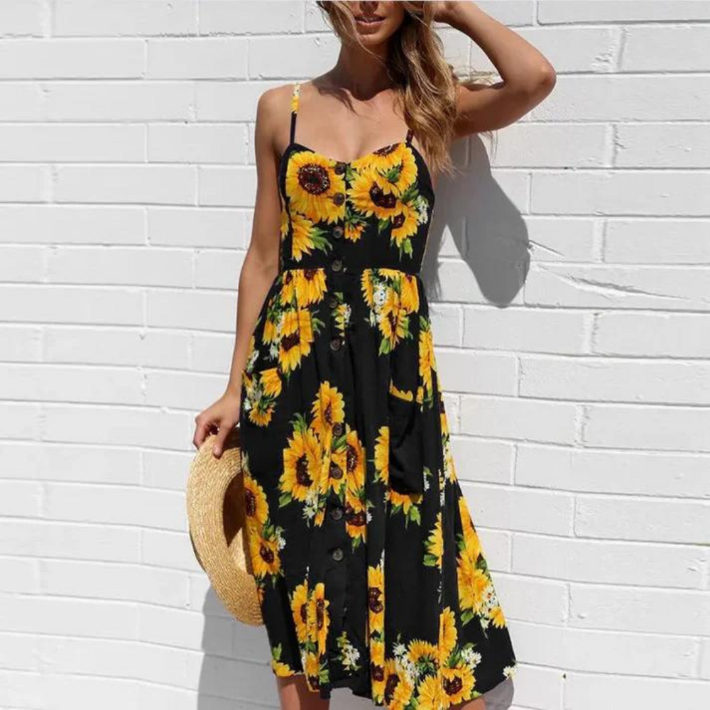 Frau in schwarzem Sonnenblumenkleid vor weißer Ziegelwand, hält Strohhut. Sommermode, Blumenkleid, Damenbekleidung, Freizeitlook.