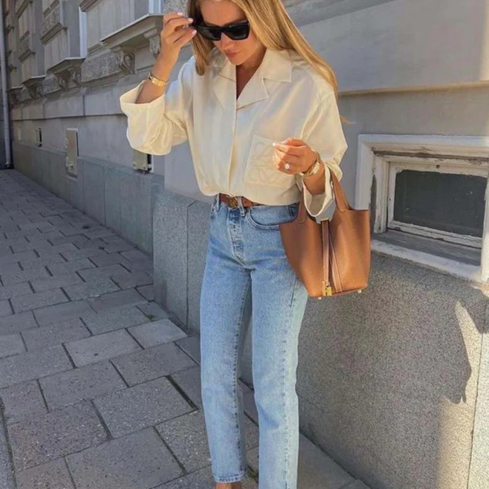Frau in weißer Bluse und blauer Jeans, trägt braune Handtasche und Sonnenbrille, steht auf städtischem Gehweg. Streetstyle, Mode, lässig.