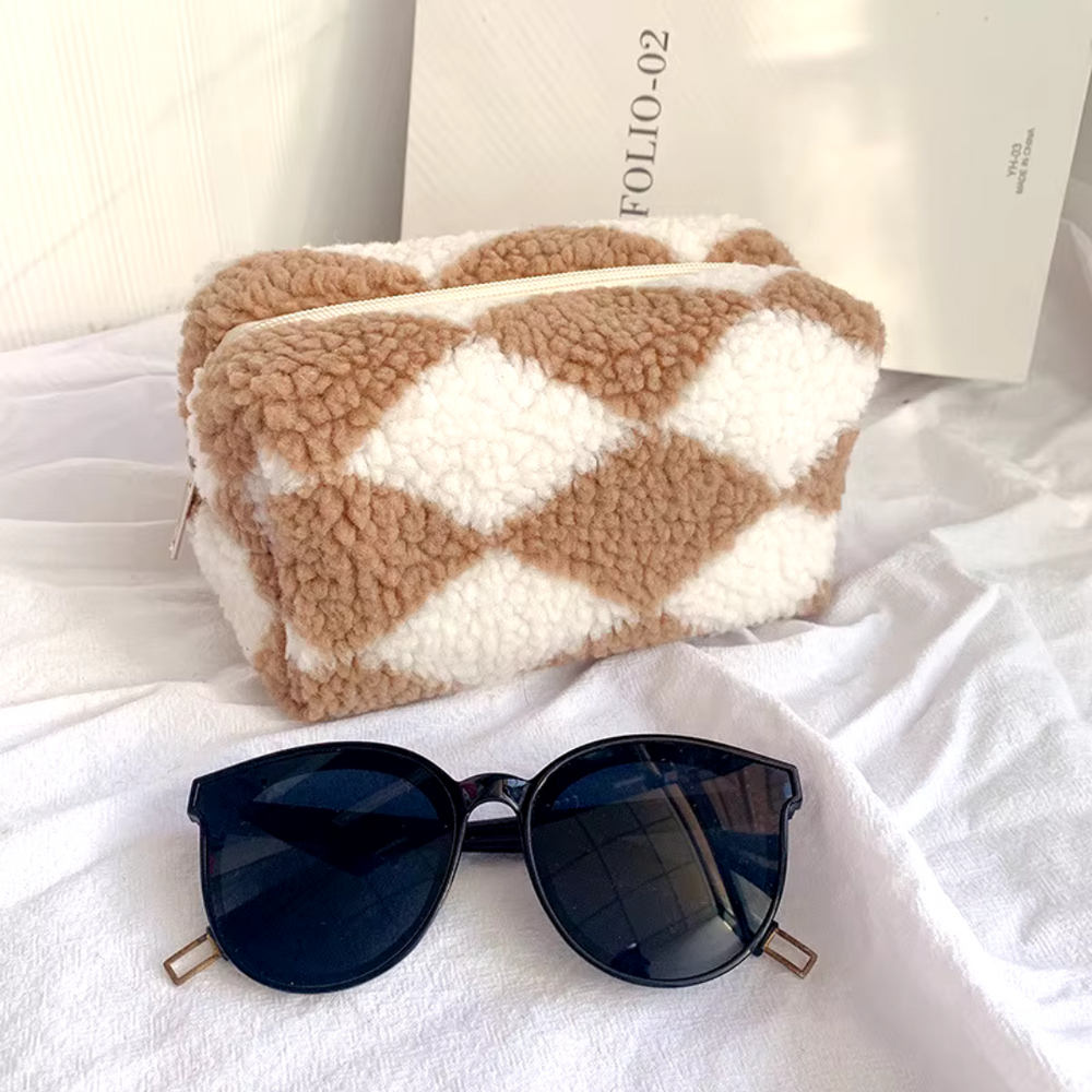 Braune und weiße Plüsch-Kosmetiktasche mit Rautenmuster, Sonnenbrille im Vordergrund, auf weißem Stoff. Trendige Accessoires, stilvoll und modern.