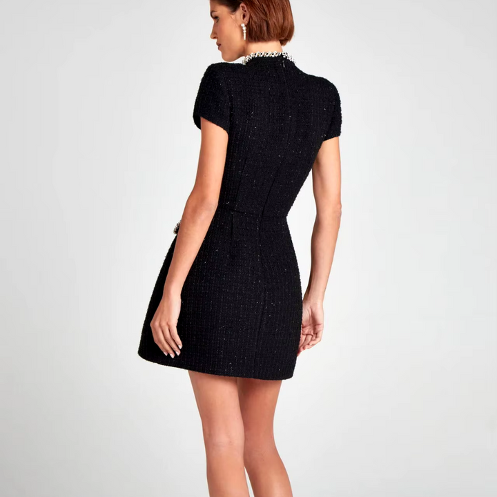 Schwarzes, kurzes Tweed-Kleid mit kurzen Ärmeln und Perlenkragen, getragen von einer Frau. Elegantes Design, ideal für Abendmode und besondere Anlässe.