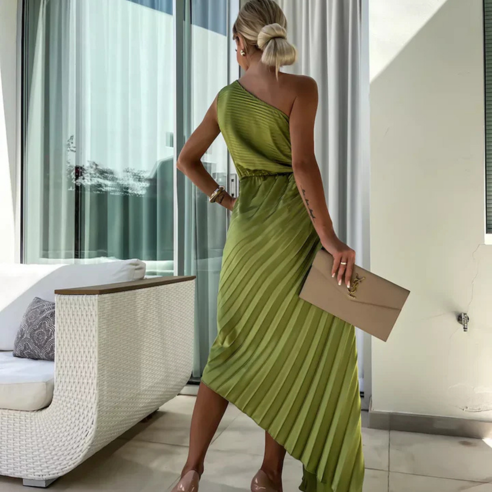 Frau in elegantem, einseitigem, grünem Plisseekleid, hält beigefarbene Clutch, steht vor moderner Terrasse mit Glasfenstern.