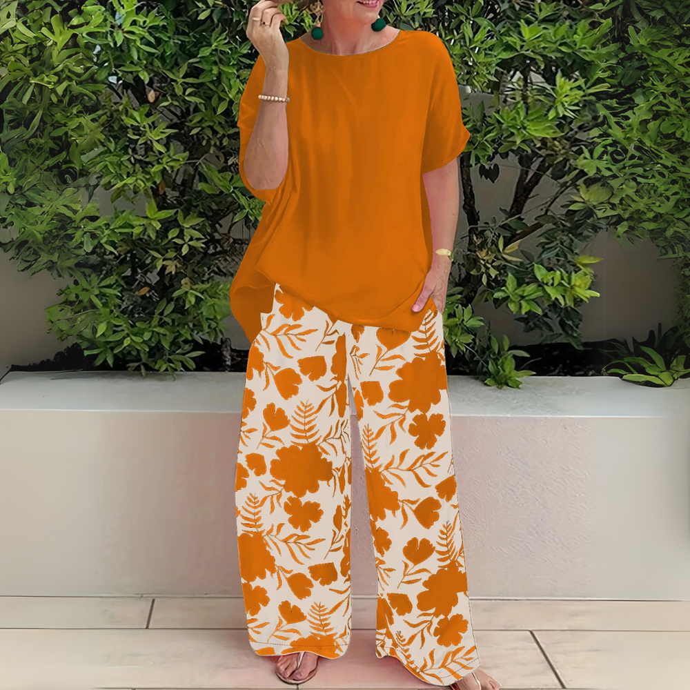 Frau in orangefarbenem Oberteil und geblümter Hose vor grüner Hecke. Sommermode, lässiger Stil, Damenbekleidung, modisches Outfit.