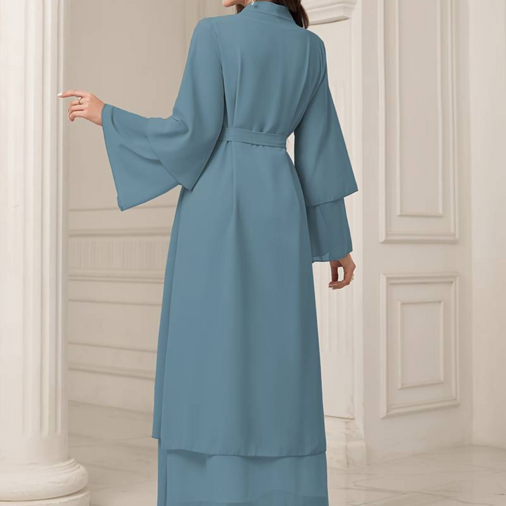 Frau in langem, blauem Kimono-Kleid mit weiten Ärmeln, steht in elegantem, weißem Interieur. Mode, Damenbekleidung, elegantes Design.