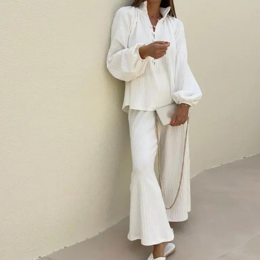 Frau in weißem, lockerem Zweiteiler mit weiten Ärmeln und Hose, hält goldene Kette. Elegante, minimalistische Mode vor beigem Hintergrund.