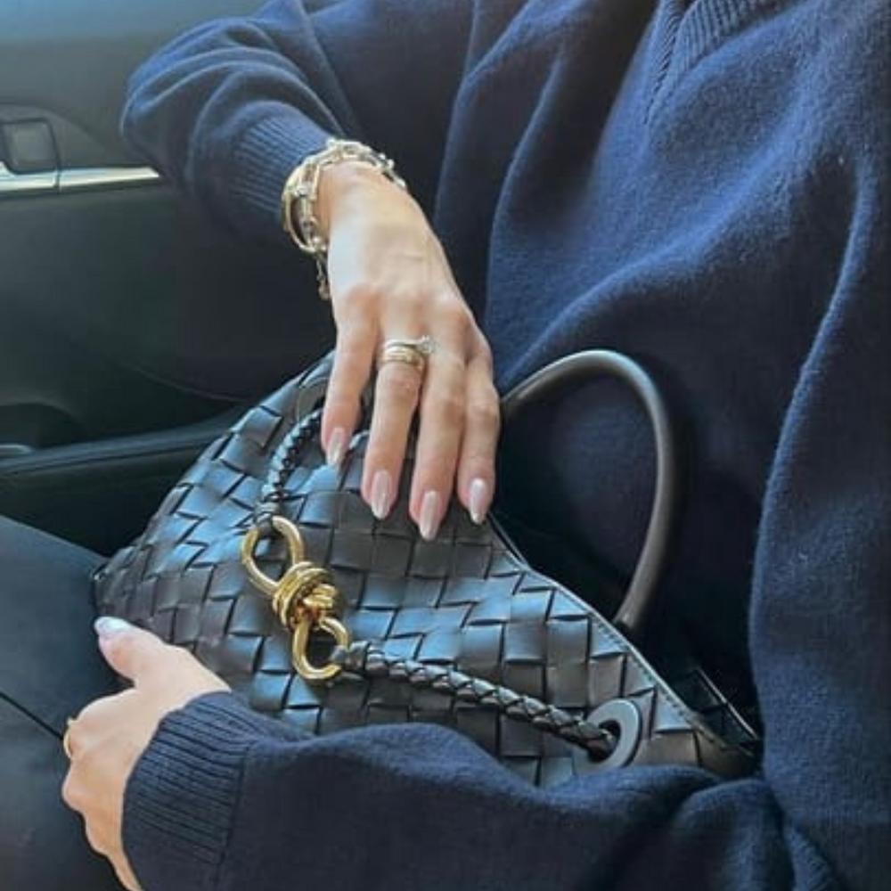 Frau im Auto mit schwarzer, geflochtener Designer-Handtasche und goldener Verzierung, elegante Maniküre, blauer Pullover, luxuriöses Accessoire.