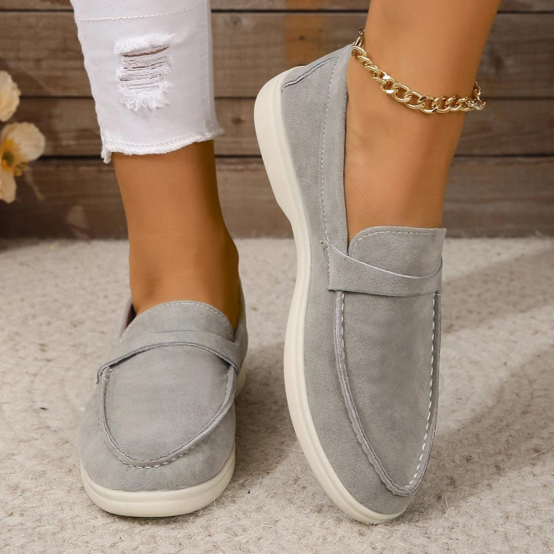 Graue Damen-Loafer aus Wildleder, kombiniert mit weißen, zerrissenen Jeans. Modische, bequeme Schuhe für den Alltag. Eleganter Look mit goldener Fußkette.
