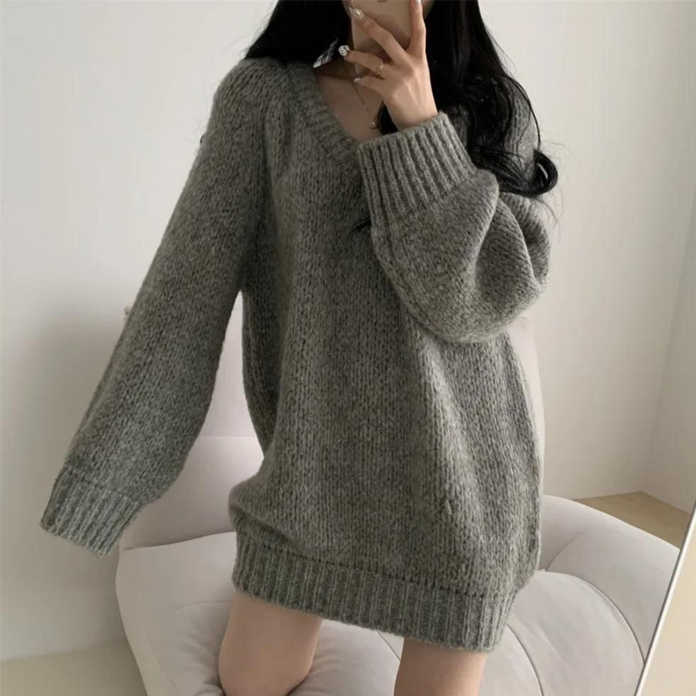 Frau in grauem Oversize-Strickpullover, gemütlich und modisch, sitzt auf einem weißen Sofa. Wintermode, lässiger Stil, bequeme Kleidung.
