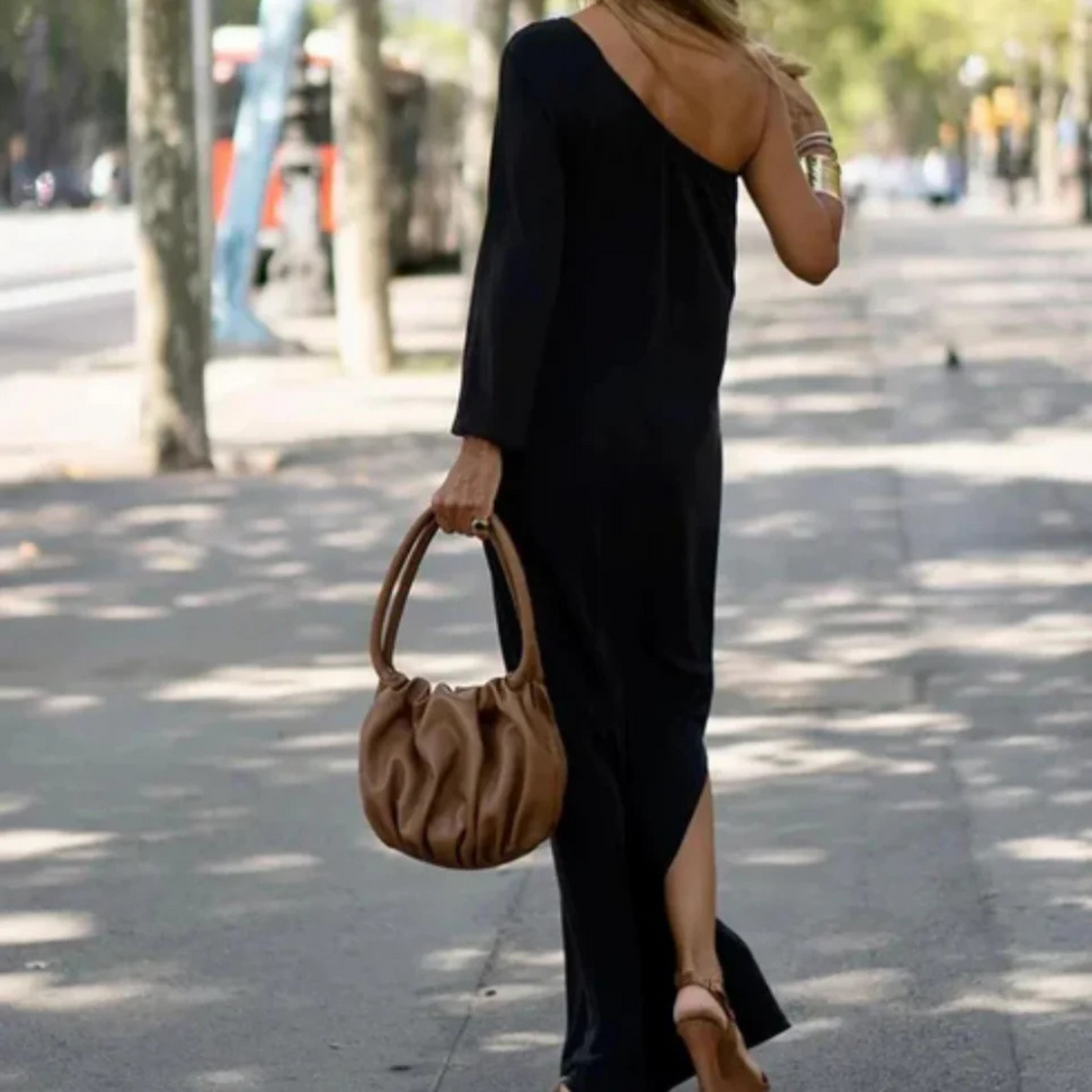 Frau in schwarzem, schulterfreiem Kleid mit brauner Handtasche spaziert auf sonniger Straße. Elegante Mode, Sommeroutfit, Stadtbummel.