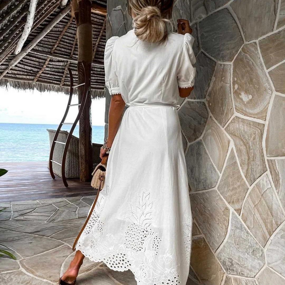 Frau in weißem Sommerkleid mit Ananas-Stickerei steht auf Terrasse mit Meerblick. Elegantes Urlaubsoutfit, tropische Kulisse, Strandmode.