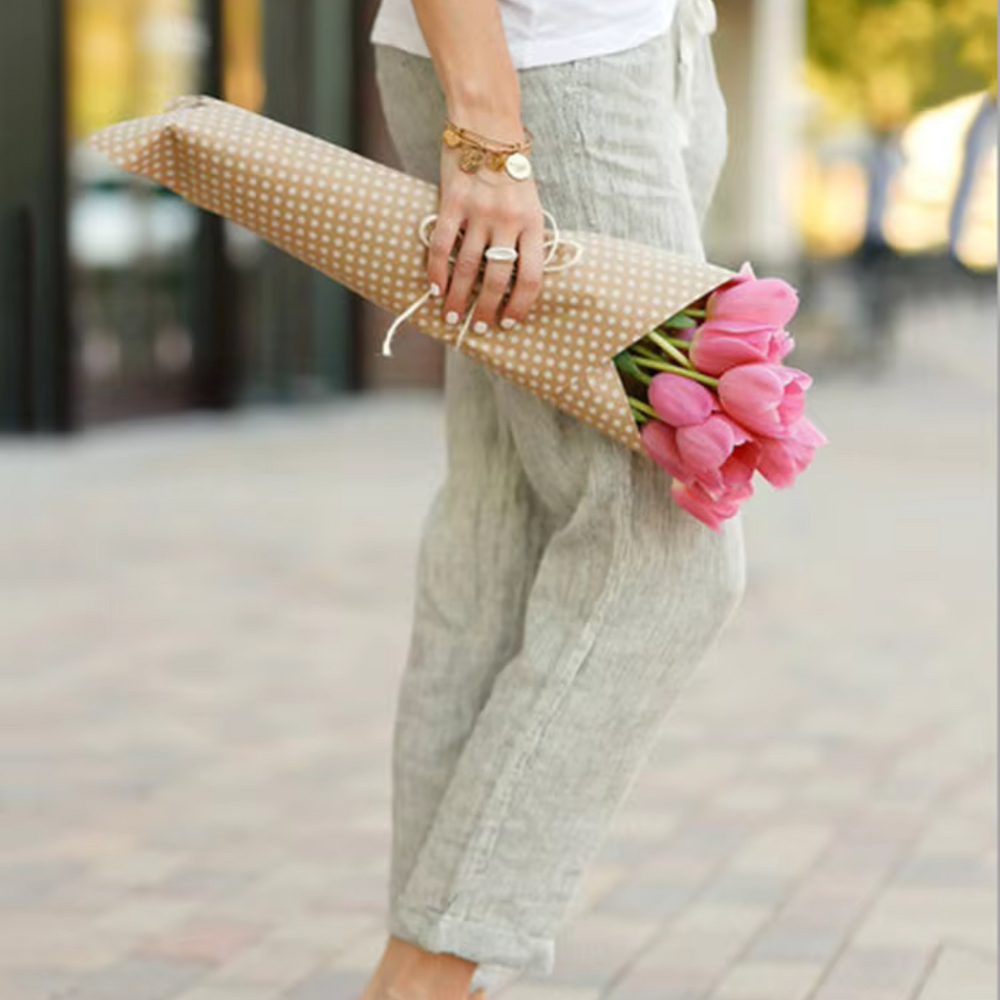 Frau hält Strauß rosa Tulpen in gepunktetem Papier, trägt lässige Leinenhose und Armbänder. Frühlingsblumen, Mode, Straßenstil.