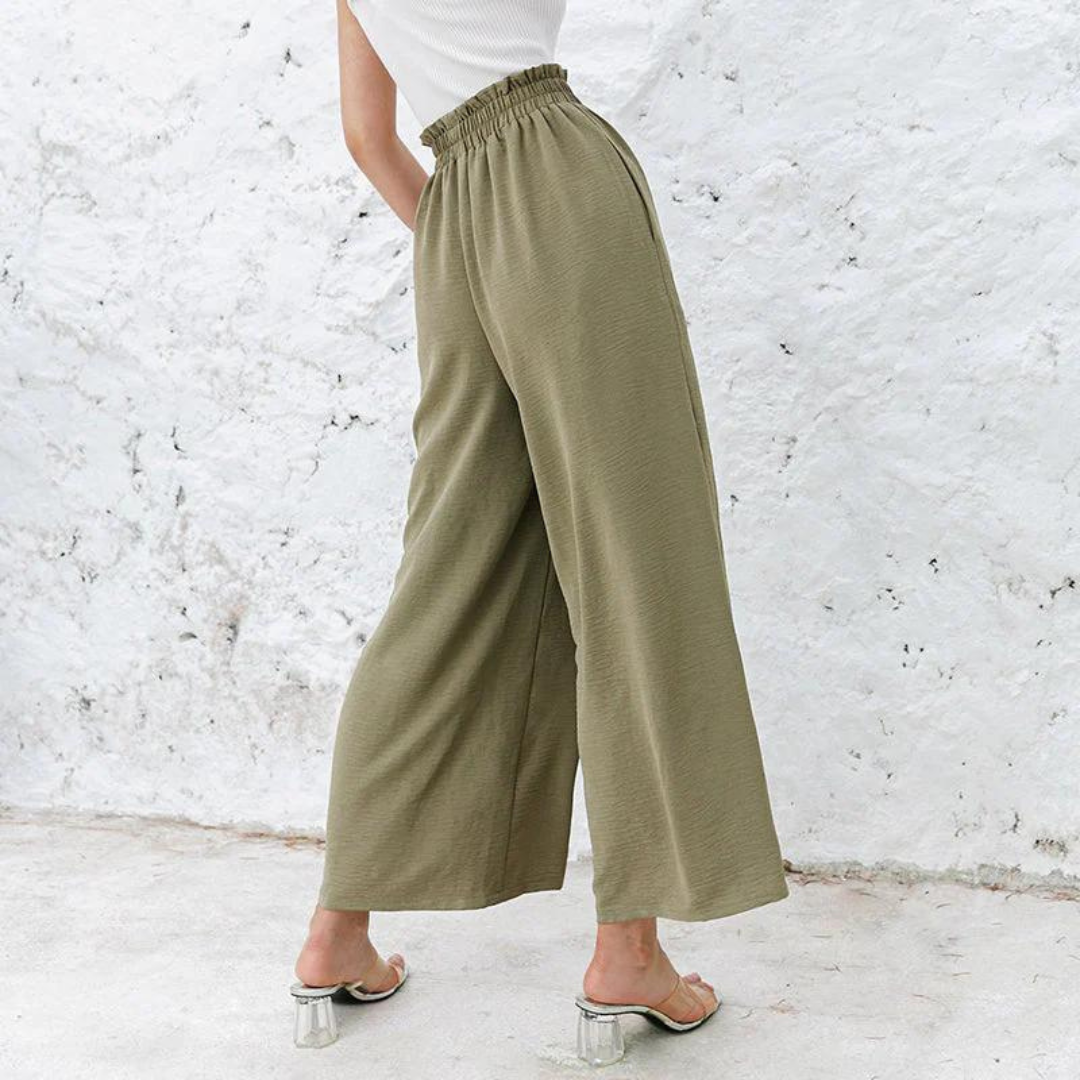Frau in olivgrüner, weiter Hose mit Gummibund und transparenten Absatzsandalen vor weißer Wand. Modetrend, Sommeroutfit, Damenbekleidung.