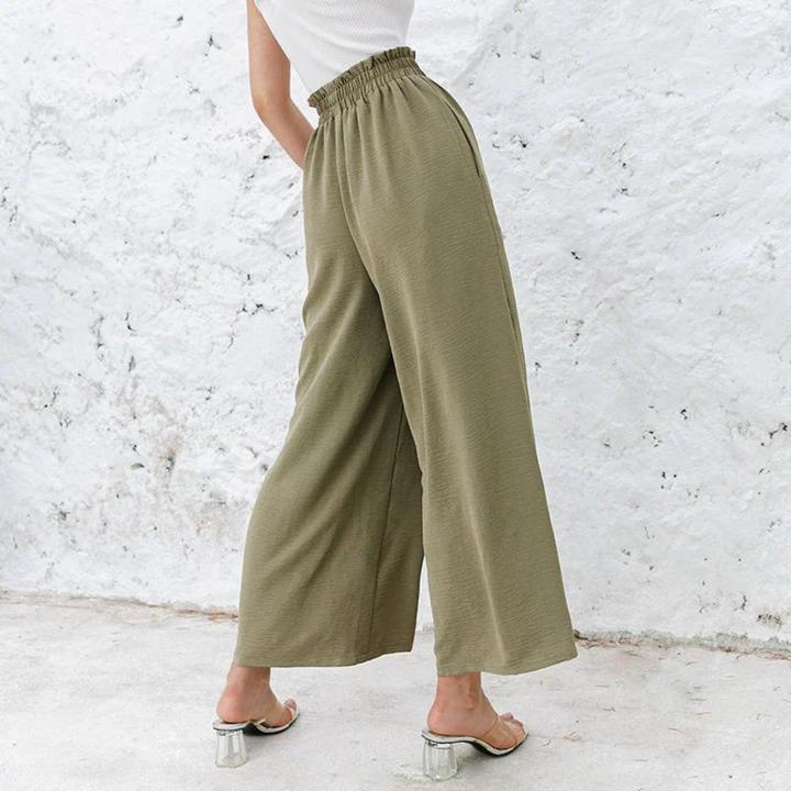 Frau in olivgrüner, weiter Hose mit Gummibund und transparenten Absatzsandalen vor weißer Wand. Modetrend, Sommeroutfit, Damenbekleidung.