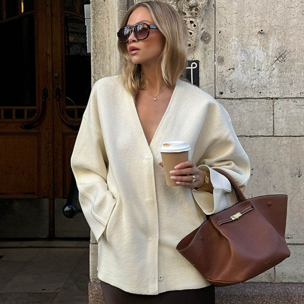 Frau in beiger Jacke mit Sonnenbrille hält Kaffeebecher und braune Ledertasche, steht vor historischer Gebäudewand. Fashion, Streetstyle, Herbstmode.