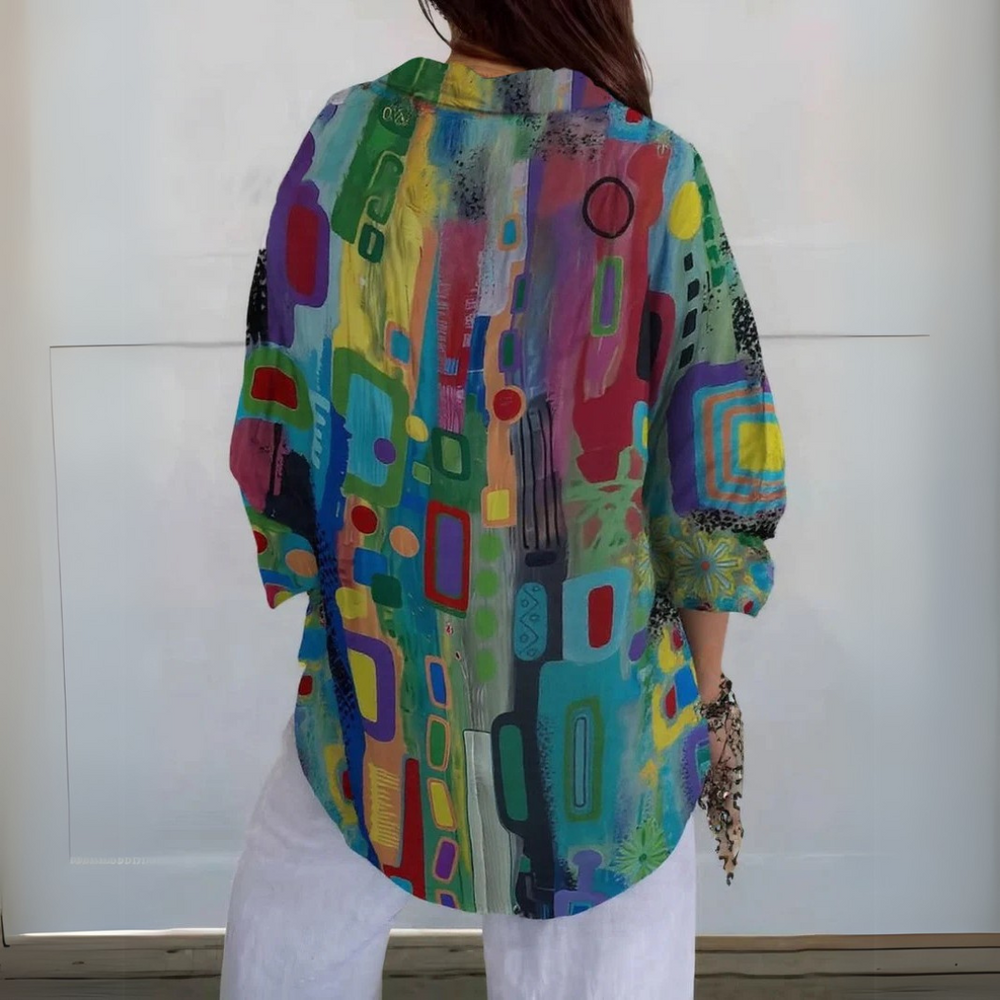 Frau trägt bunte, abstrakte Kunstbluse mit geometrischen Mustern, kombiniert mit weißen Hosen. Modetrend, farbenfrohe Damenbekleidung, lässiger Stil.