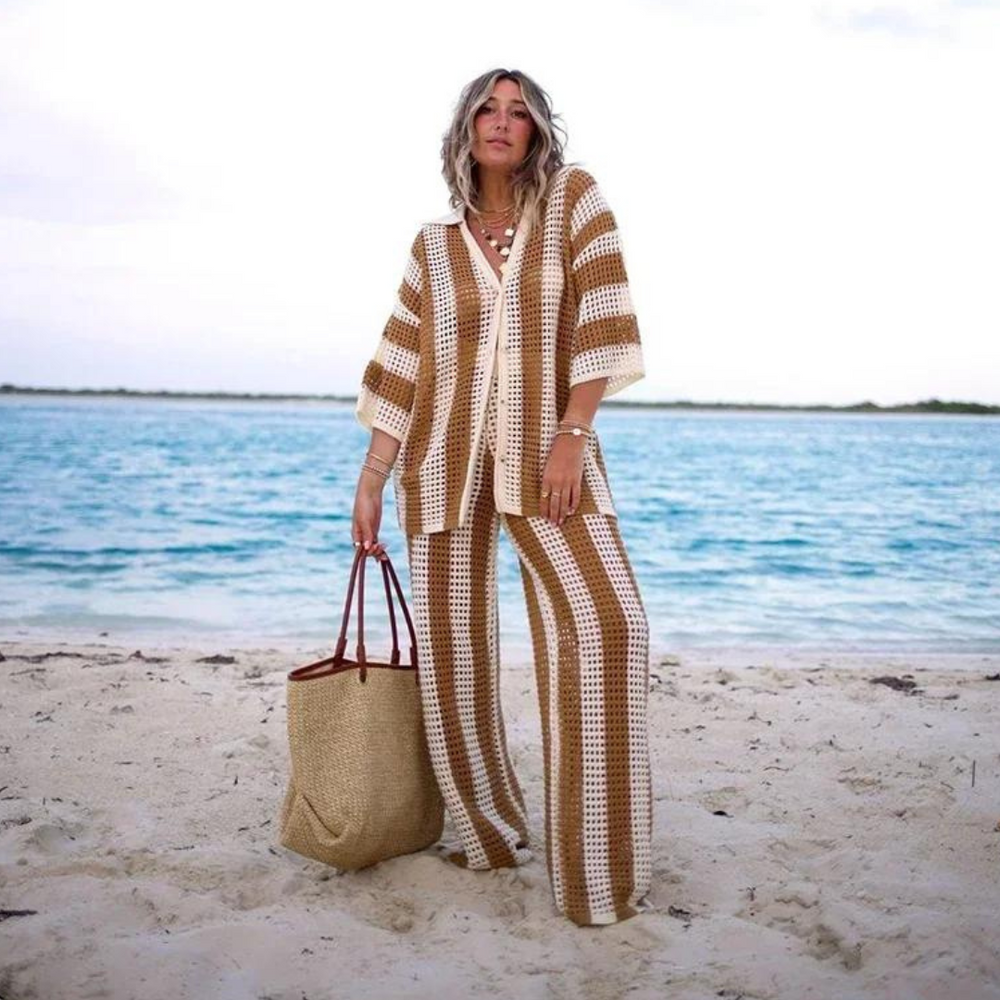 Frau am Strand in gestreiftem Häkelanzug, hält eine Strohtasche. Sommermode, Strandkleidung, Häkeltrend, entspannter Look, Küstenhintergrund.