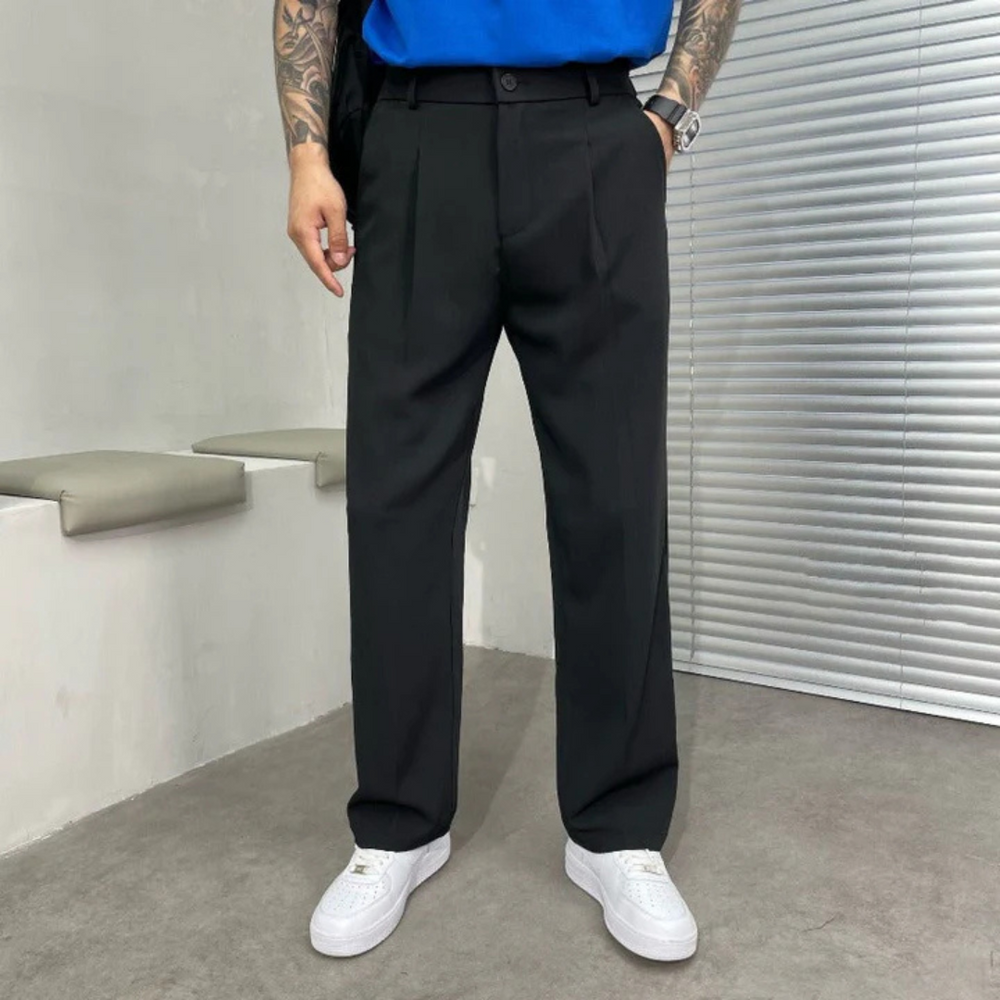 Mann in schwarzer Anzughose und weißen Sneakers, steht vor einer grauen Wand mit Jalousie. Moderne Herrenmode, lässiger Business-Look.
