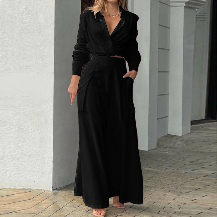 Frau in eleganter schwarzer Bluse und weiter Hose, steht barfuß auf gepflastertem Boden vor weißer Wand. Modetrend, Damenmode, schickes Outfit.