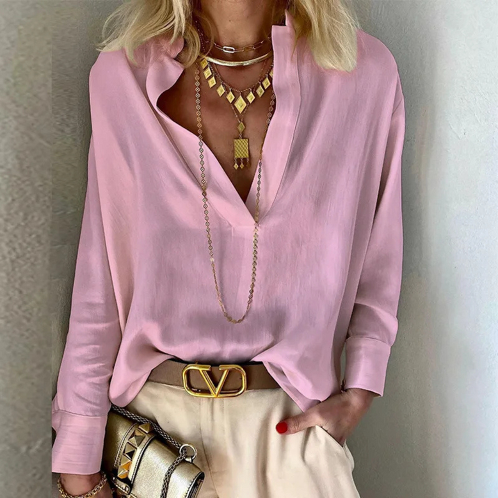 Blonde Frau in rosa Bluse mit tiefem Ausschnitt, goldenen Halsketten und Gürtel mit Designer-Schnalle, hält goldene Clutch. Elegante Mode, Schmuck.