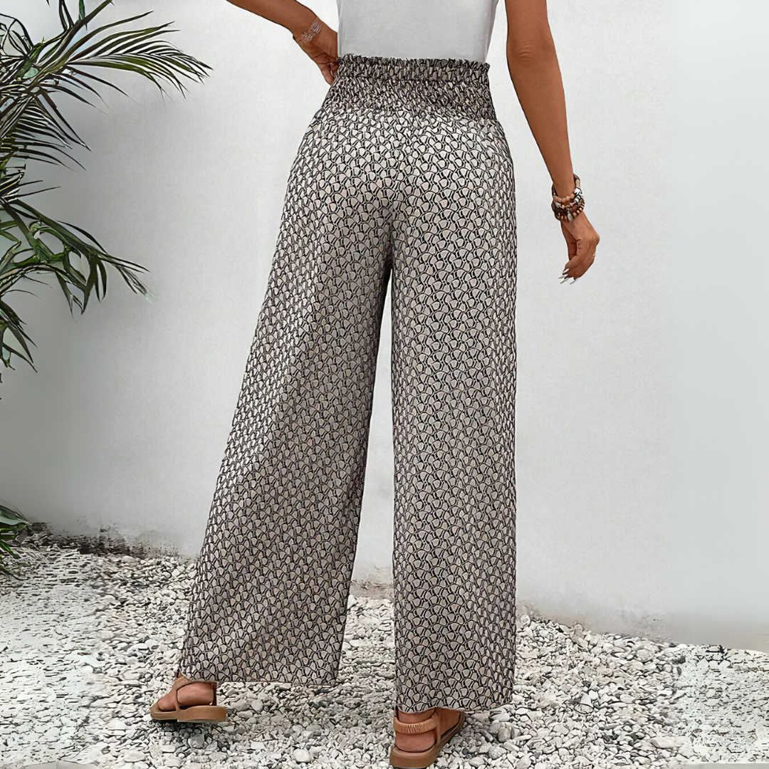 Frau in gemusterten, weiten Hosen und weißem Top, steht auf Kiesboden neben grüner Pflanze. Modetrend, Sommeroutfit, Damenbekleidung.