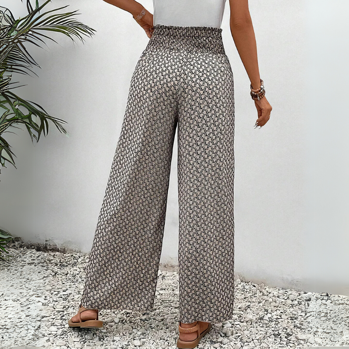 Frau in gemusterten, weiten Hosen und weißem Top, steht auf Kiesboden neben grüner Pflanze. Modetrend, Sommeroutfit, Damenbekleidung.