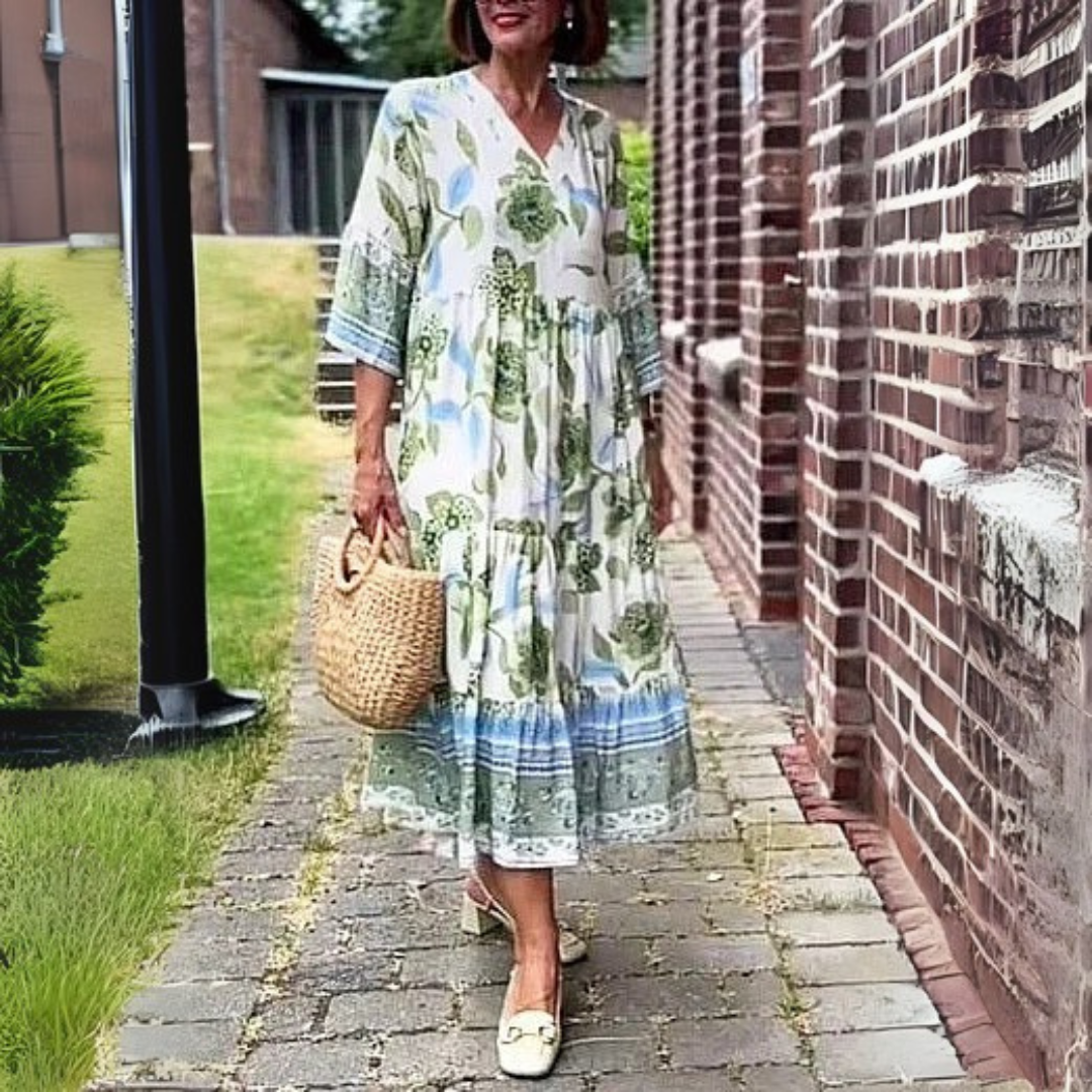 Frau in geblümtem Sommerkleid und Strohtasche spaziert auf gepflastertem Weg neben Backsteinmauer. Elegante Mode, Sommeroutfit, Freizeitkleidung.