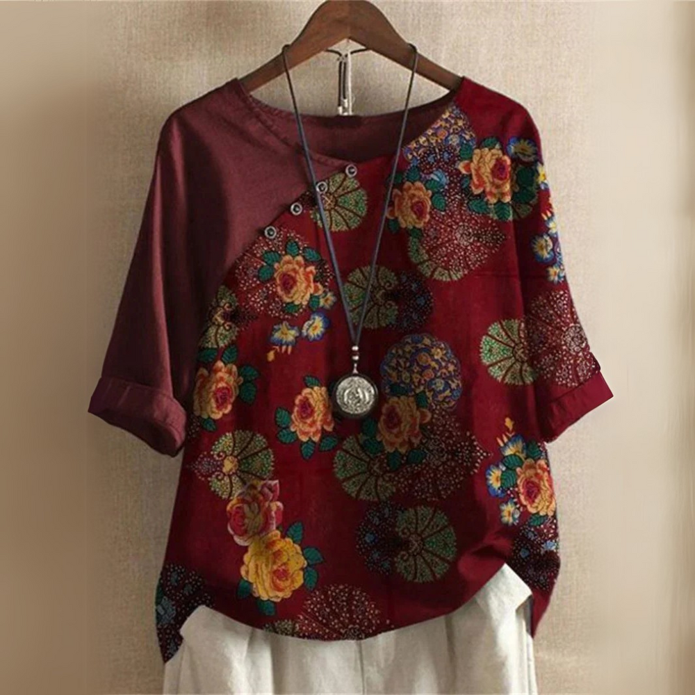 Buntes Damenoberteil mit Blumenmuster, asymmetrischem Design und Knopfleiste, hängend auf Holzkleiderbügel. Modisches, florales Shirt.