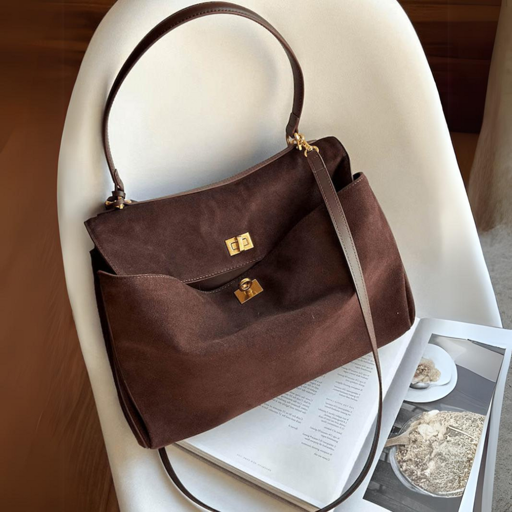 Braune Wildleder-Handtasche mit goldenen Verschlüssen auf einem weißen Stuhl, neben einem aufgeschlagenen Magazin. Elegante Damenmode, Accessoire.