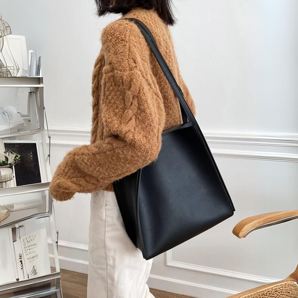 Frau mit schwarzer Ledertasche, braunem Strickpullover und weißer Hose in modernem Raum. Modetrend, Damenaccessoire, stilvoller Look.