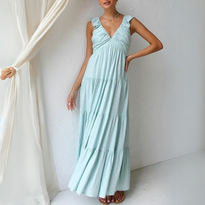 Frau in langem, hellblauem Sommerkleid mit Rüschenärmeln steht vor weißer Wand. Elegantes Maxikleid, modisches Design, luftiger Stoff.