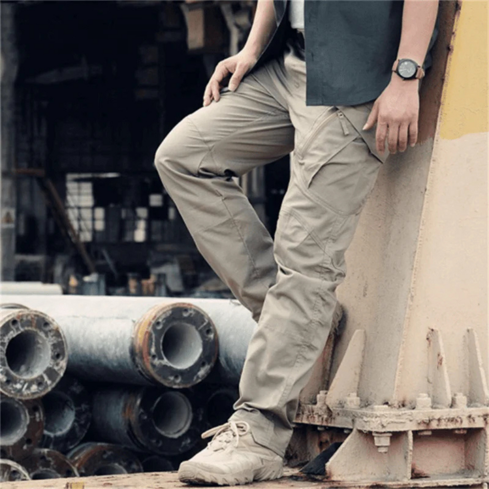 Mann in beigen taktischen Hosen und schwarzem Hemd lehnt an Industrieanlage. Outdoor-Mode, robuste Kleidung, urbaner Stil.