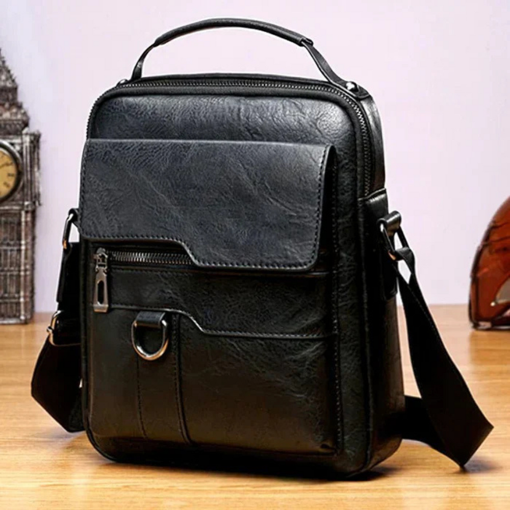 Schwarze Leder-Umhängetasche auf Holztisch, mit Reißverschluss, Fronttasche und verstellbarem Schultergurt. Ideal für Herrenmode und Business.