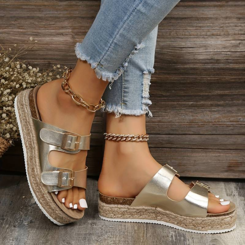 Goldene Plateau-Sandalen mit doppelten Schnallen an Frauenfüßen, kombiniert mit zerrissenen Jeans und Fußkettchen, vor rustikalem Holzhintergrund.
