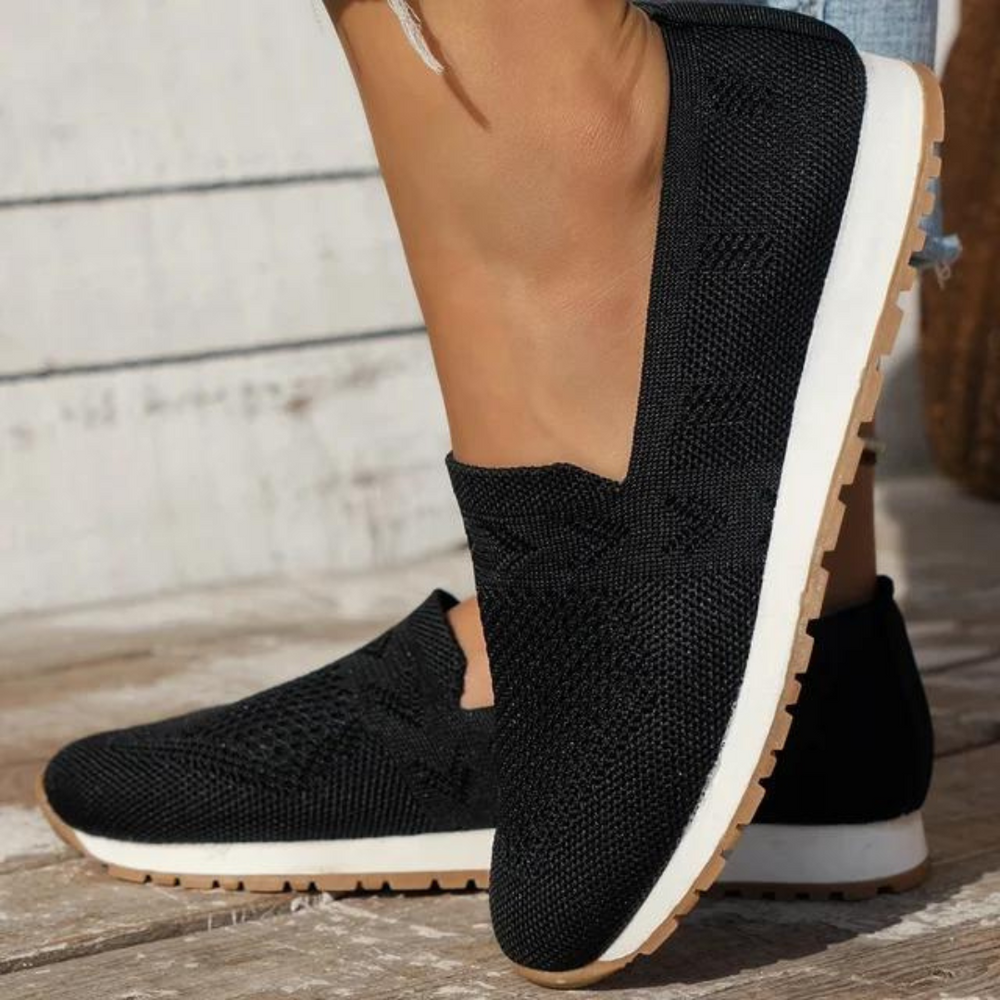 Schwarze Damen-Sneaker aus Strickmaterial mit weißer Sohle und braunem Profil, getragen auf Holzboden. Bequeme, modische Freizeitschuhe.