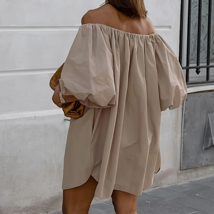 Frau in beigem schulterfreiem Kleid mit Puffärmeln, hält goldene Clutch. Modetrend, eleganter Streetstyle, Sommermode.