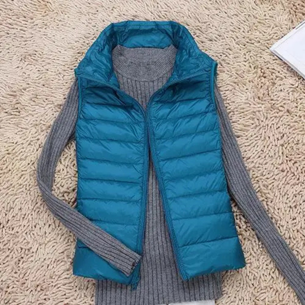 Blaue Steppweste auf grauem Strickpullover, liegend auf beigem Teppich. Modische Damenbekleidung, Herbstmode, warme Oberbekleidung.