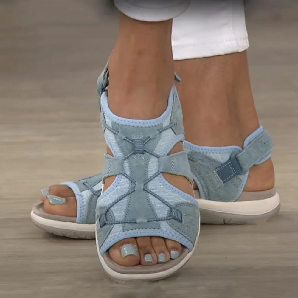 Damenfüße in hellblauen, offenen Sandalen mit Klettverschluss, getragen auf hellem Holzboden. Modische Sommerschuhe, bequem und stilvoll.